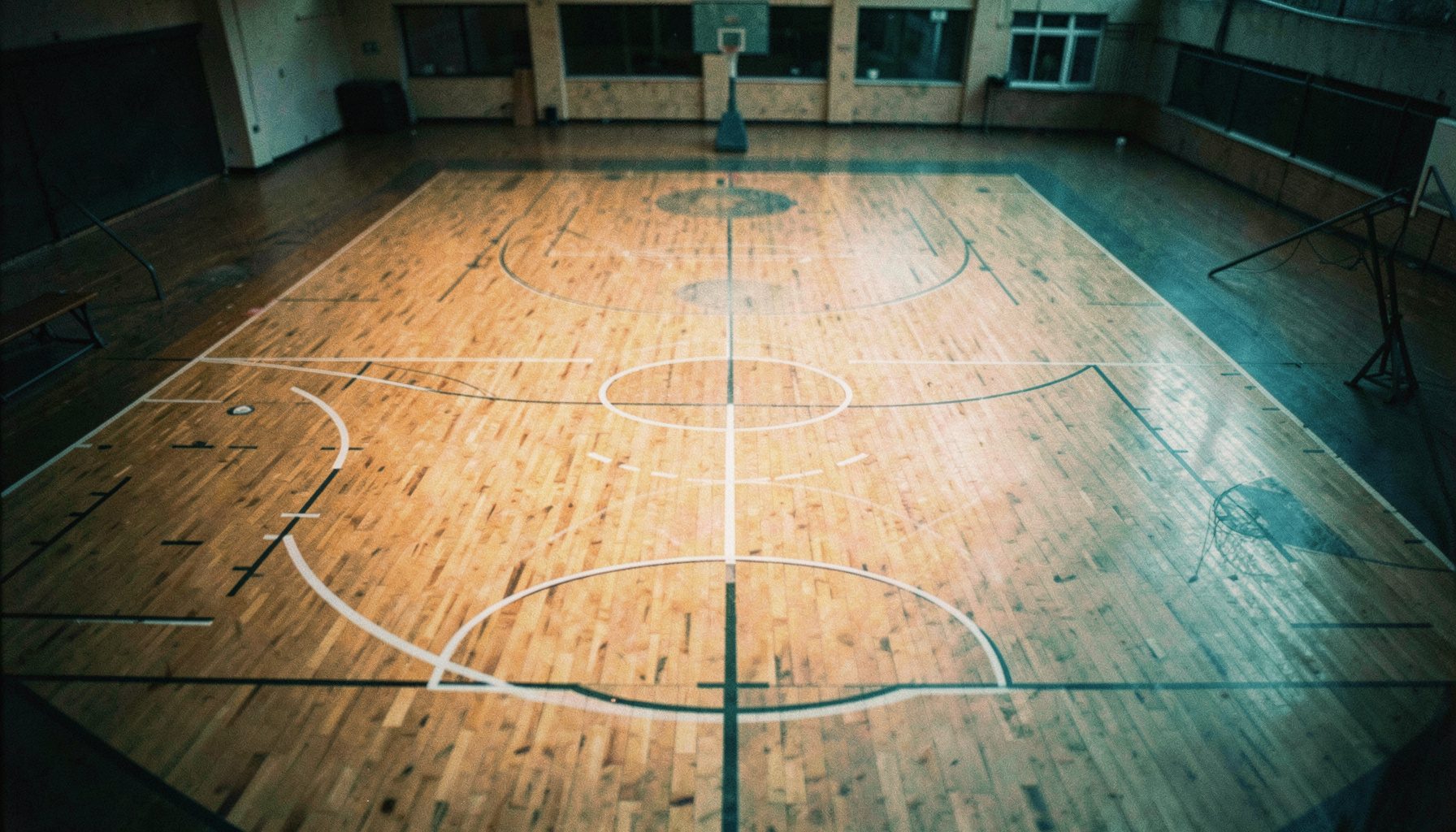 Turuncu ve lacivert tonlarında, bir basketbol sahasının havadan geniş açılı görünümü; parke zemin ve pota silüeti
