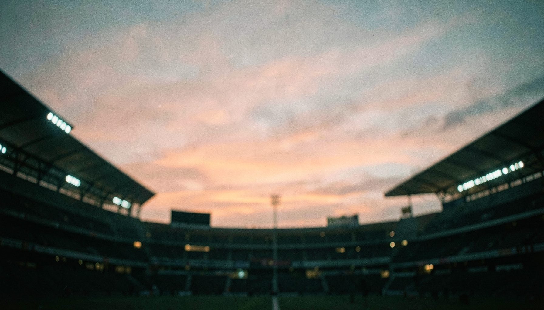 Geniş açılı, yumuşak odaklı bir stadyum silüeti; turuncu ve lacivert gökyüzü tonlarıyla sakin bir atmosfer.